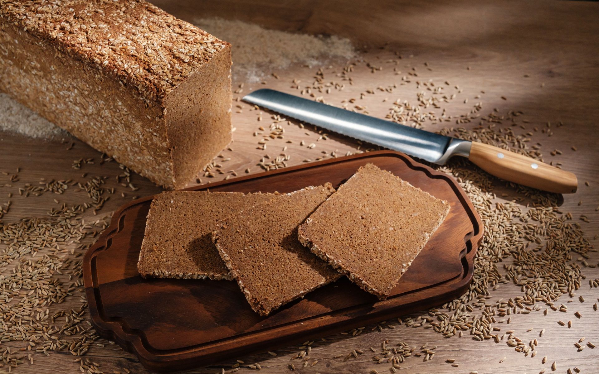 Whole-grain loaf with seed crust, three slices on wooden board, bread knife and scattered grains