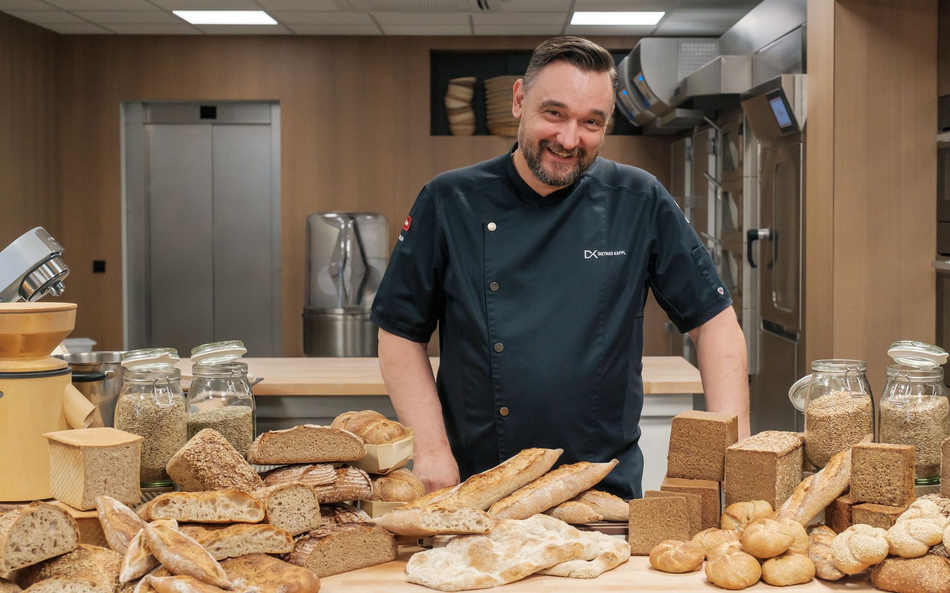 Assorted whole-grain loaves, baguettes and rolls on a wooden table, jars of grains, baker in professional kitchen