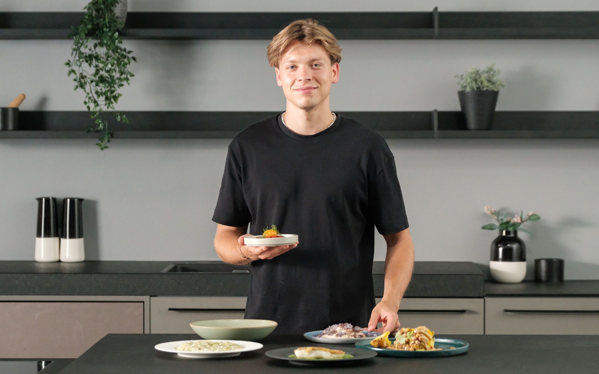 Chef presenting plated fine-dining dishes in a modern kitchen, several colorful plates on the counter